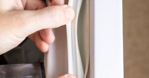 Two hands pulling a white gasket off the side of a refrigerator’s door, warping and leaving a gap in between.