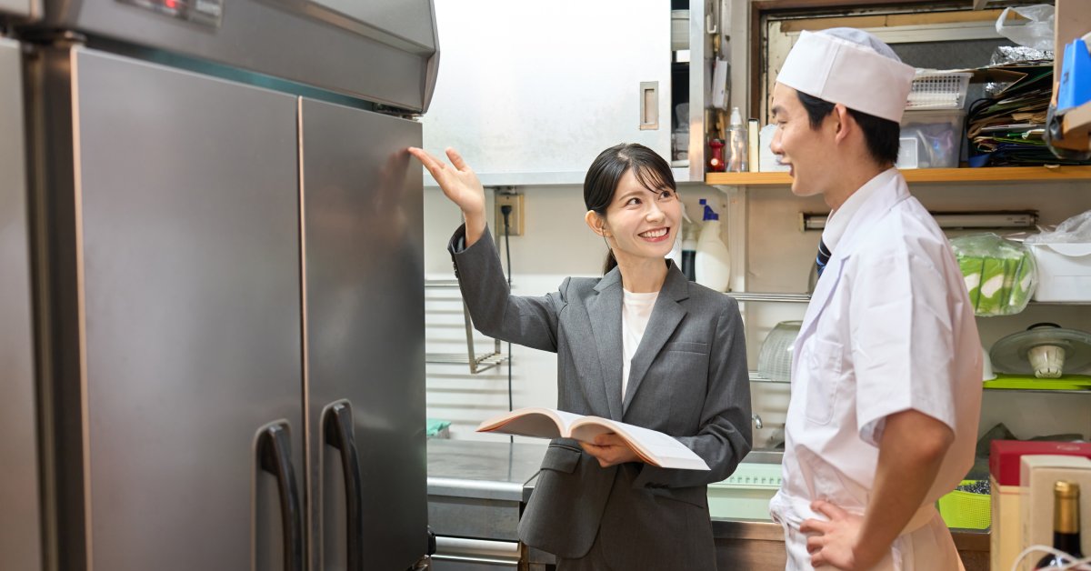 A woman in a suit holding up a hand next to a freezer and smiling at a chef standing next to her inside a kitchen.
