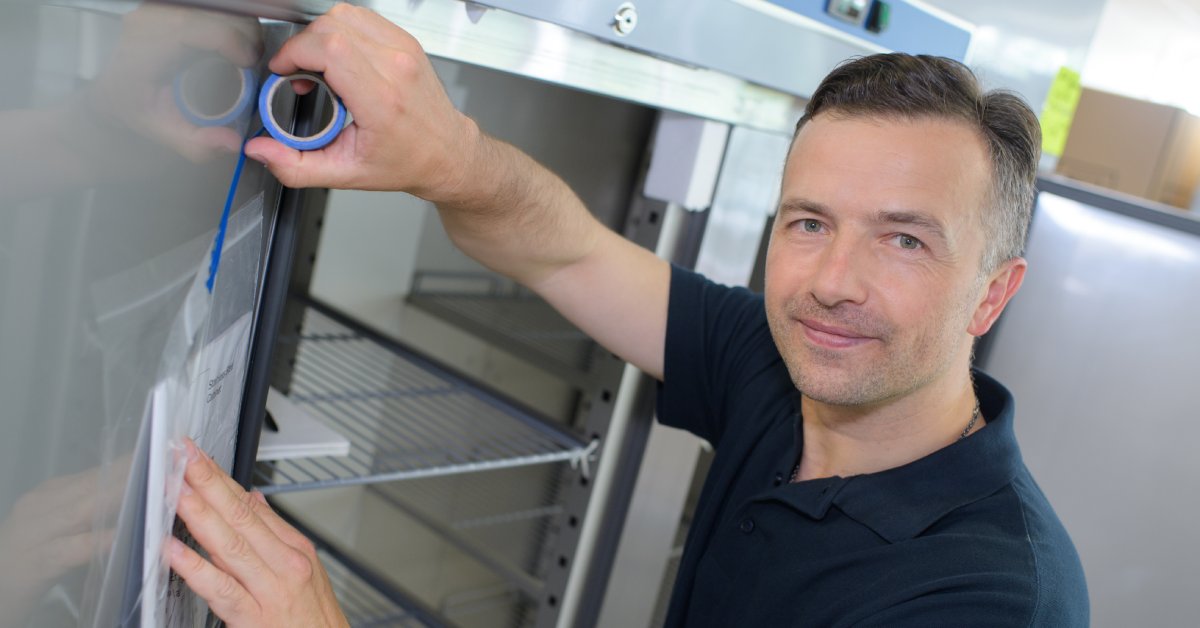 Man taping a piece of paper inside a plastic cover to the interior of a refrigerator’s door with black rubber gaskets.
