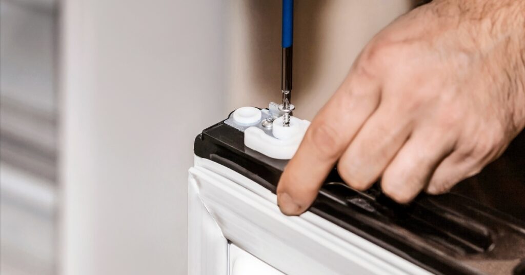A close-up of a person using a screwdriver to fix the top part of a refrigerator door. The door has a white gasket.