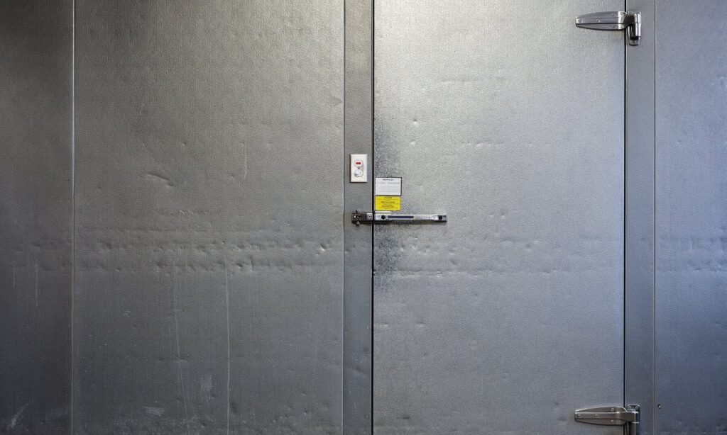 A metal freezer door with two metal hinges has two locks to secure it. Once lock is on the handle and the other is on the top of the door frame.