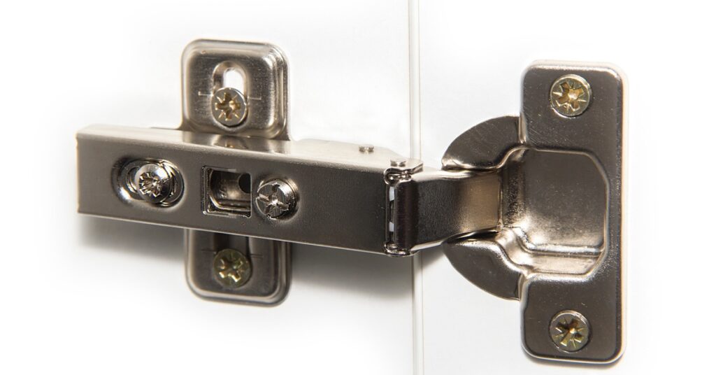 Close-up to a large metal hinge with multiple Philips screws and a chrome finish placed on a white metal door.