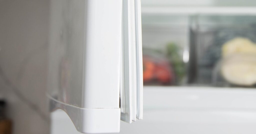 A close-up of a white gasket with a separated corner from a white freezer door, with blurred food in the background.