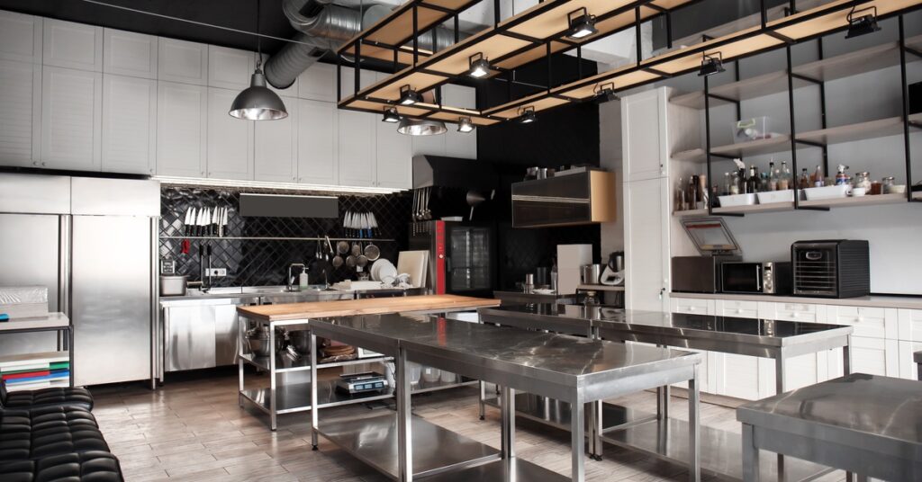 A large, empty restaurant kitchen with stainless steel counters and multiple overhead lights on a wooden structure