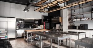 A large, empty restaurant kitchen with stainless steel counters and multiple overhead lights on a wooden structure