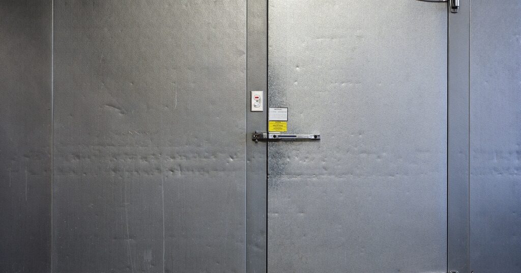 A close-up of a large freezer door, with two large metal hinges and two safety locks on the top and the side.