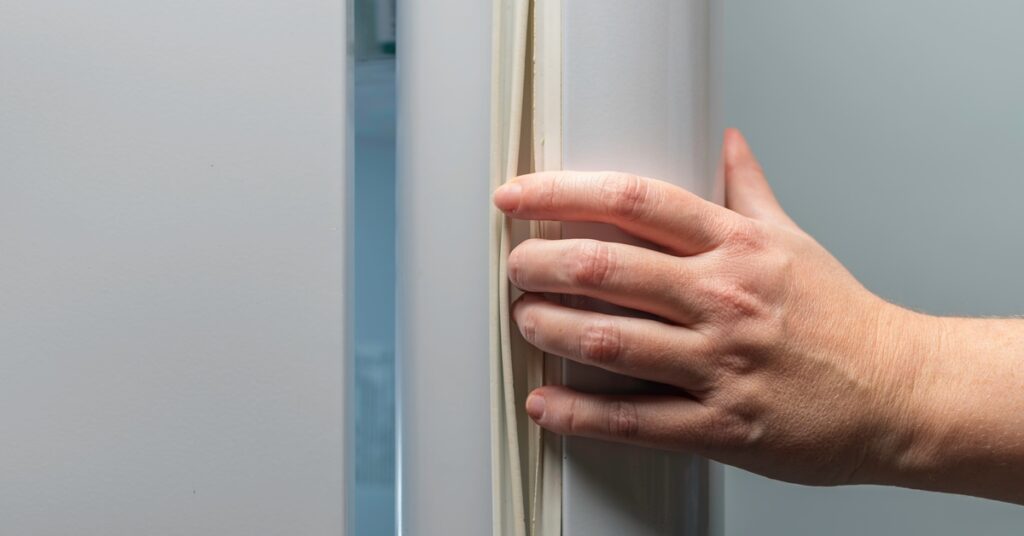 A close-up of a hand sticking its fingers between a white rubber gasket and an open, large freezer door.