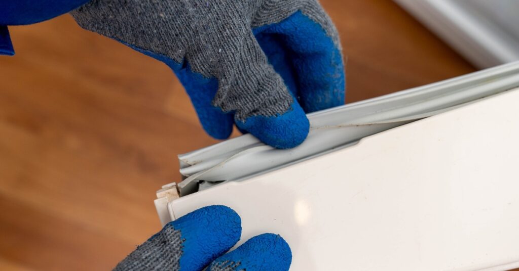A close-up of two hands wearing grey gloves with blue tips, inspecting the white rubber gasket placed on a freezer door.