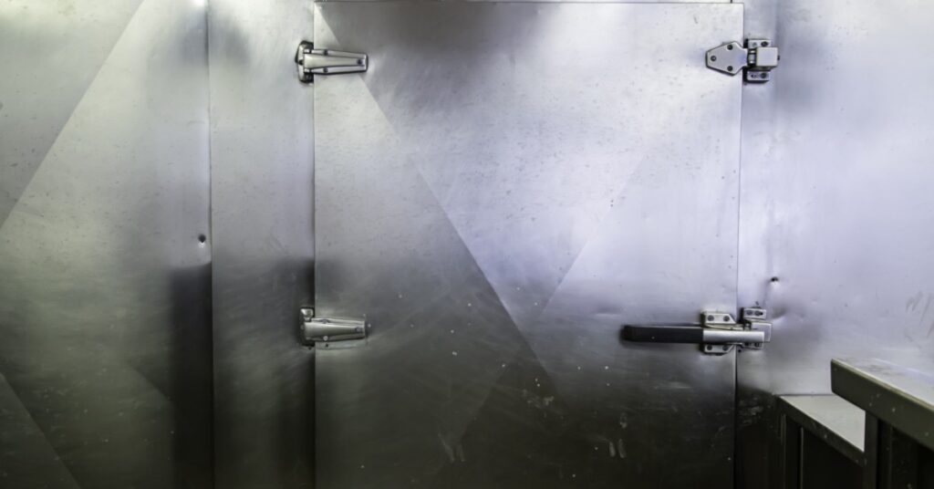 A large metal door with multiple hinges and a safety lock, placed on a metal wall, next to a stainless steel counter.