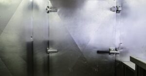 A large metal door with multiple hinges and a safety lock, placed on a metal wall, next to a stainless steel counter.
