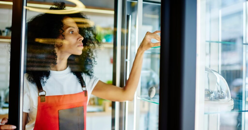 A woman wearing an orange apron holding a large glass freezer door with a black trim open, sticking her hand inside.