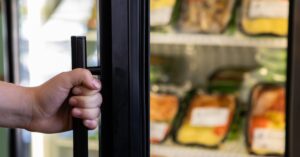 A close-up of a hand holding the door of a large refrigerator with black, thick gaskets, with food in the background.