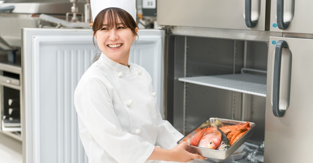 A woman wearing a white coat and a white chef hat holding a metal tray with a fish next to the open door of a freezer.