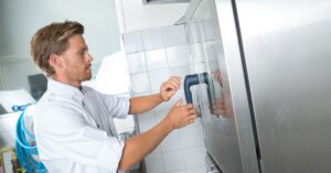 A man in a white uniform touching a handle attached to a stainless-steel refrigerator door in a commercial kitchen.