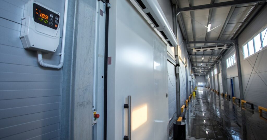 A large commercial refrigerator door with a black gasket, next to a digital thermometer on the wall in a long hallway.
