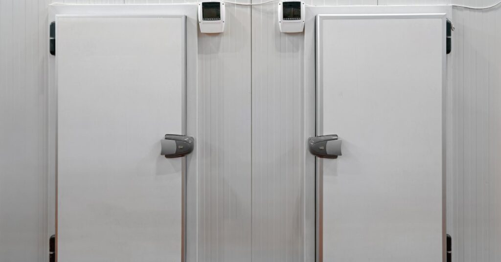 Two white commercial freezer doors next to each other, with grey handles and black hinges, in a room with red floors.