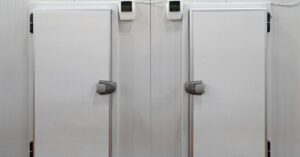 Two white commercial freezer doors next to each other, with grey handles and black hinges, in a room with red floors.