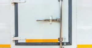 A close-up of a large white door with a metal handle bar crossing from top to bottom and large white hinges.