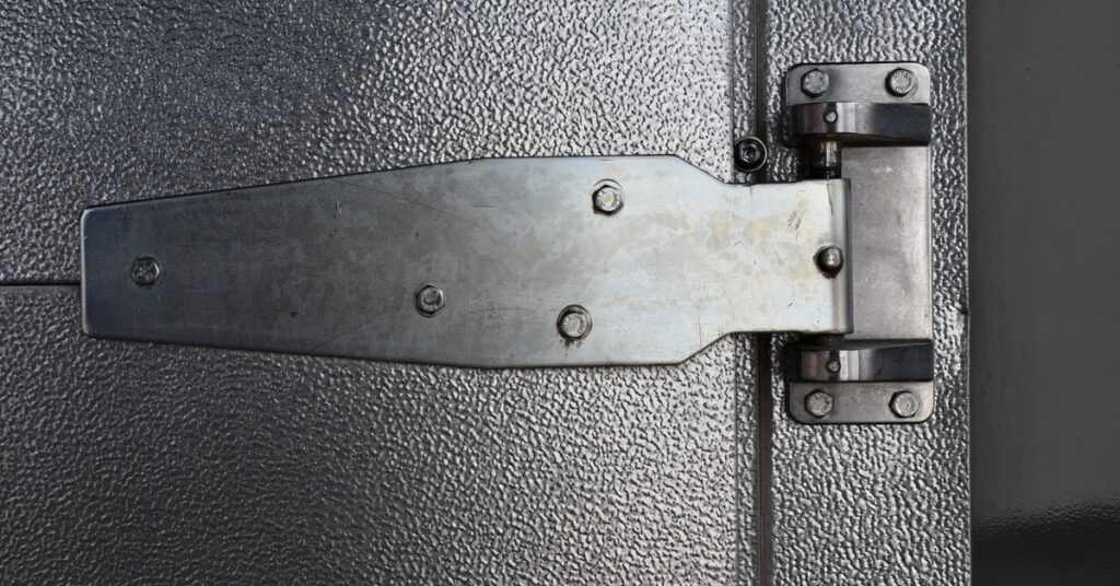 A close-up of a large metal hinge with small screws, placed on the edge of a metal, grey door with a shiny finish.