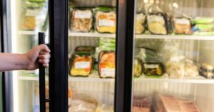 A hand holding a commercial freezer door with black gaskets open with multiple packed food trays inside.