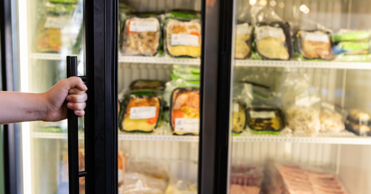 A hand holding open the glass door of a commercial refrigerator with black gaskets and multiple frozen products inside.
